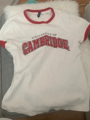 Vit Cambridge t-shirt med röd kant - Säljer en vit t-shirt med röd text och kontrasterande röda kanter vid hals och ärmslut. Klassisk och sportig look med tryck framtill. Perfekt för dig som gillar college-stil.
