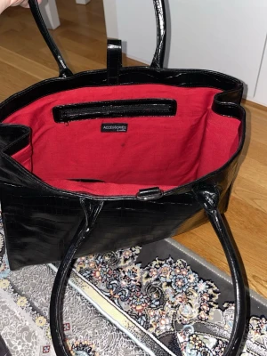 Svart croco-mönstrad handväska - Stilren svart handväska från Accessories med croco-mönstrad yta i glansigt konstläder. Väskan har två långa handtag och röd textilinsida med innerfack. Perfekt för dig som vill ha en trendig och rymlig väska med coola detaljer.