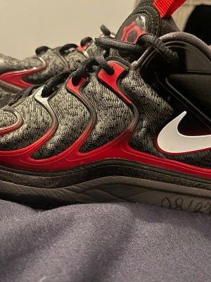 Nike KD17 svart/röd basketskor - Nike KD17 basketskor i svart, grått och rött med vita detaljer. Skorna har en stickad ovandel med röda och svarta syntetiska förstärkningar samt en vit Nike-swoosh på sidan. Designen är sportig och modern, perfekt för basket eller streetwear.