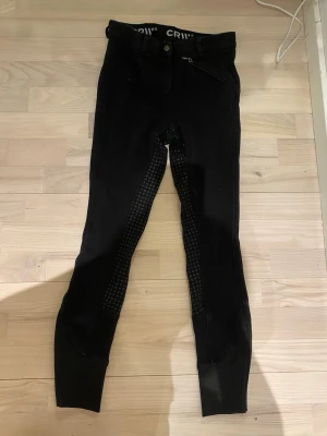 Svarta ridleggings CRW strl 160 - Svarta ridleggings från CRW med helskoning och grepp längs insidan av benen. De har en dragkedjeficka framtill och är tighta i passformen. Perfekta för dig som vill ha bekväma och funktionella leggings till ridning.