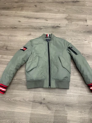 Grön bomberjacka med röda detaljer - Snygg grön bomberjacka från Tommy Hilfiger med svarta och röda detaljer. Jackan har två stora fickor framtill, en ärmficka med dragkedja och patch på ena ärmen. Ribbstickad krage, midja och ärmslut ger en klassisk look. 