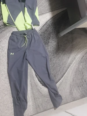 Under Armour tracksuit - Säljer ett par grå träningsbyxor från Under Armour med elastisk midja och snörning i ljusgrönt. Byxorna har en avslappnad passform och är perfekta för sport och träning. Materialet är lätt och bekvämt, idealiskt för aktiva dagar.