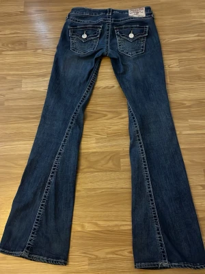 Mörkblå bootcut jeans från true religion  - Säljer ett par mörkblå bootcut jeans från true religion med snygga kontrastsömmar och dekorativa bakfickor med knappar. Jeansen har låg midja och är tillverkade i klassiskt denimtyg. Perfekta för dig som gillar en avslappnad men ändå trendig look.