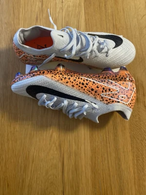 Nike Mercurial orange/vita fotbollsskor - Säljer ett par Nike Mercurial fotbollsskor i vitt och orange med svarta detaljer och leopardmönster. Skorna har en låg profil, snörning och en skinande sula med dobbar för bästa grepp på planen. Perfekta för dig som vill sticka ut på matchen!