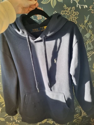 Mörkblå hoodie från Polo Ralph Lauren - Mörkblå hoodie från Polo Ralph Lauren med klassisk huva och dragsnören. Broderad vit logga på bröstet och stor känguruficka framtill. Mjuk bomullskänsla och avslappnad passform, perfekt för en chill look.