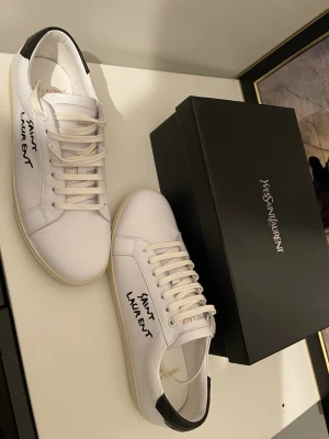 Saint Laurent vita skor - Snygga vita sneakers från Saint Laurent med svart detalj på hälen och handskriven logga på sidan. Skorna har klassisk låg profil, vita snören och ovandel i slätt läder. Perfekta för dig som gillar stilrena och exklusiva sneakers.