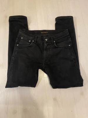 Svarta jeans från Nudie Jeans - Säljer ett par svarta jeans från Nudie Jeans med klassisk femficksdesign och raka ben. Jeansen har silverfärgade nitar och knapp, samt diskreta sömmar. Perfekta för dig som gillar en clean och stilren look.