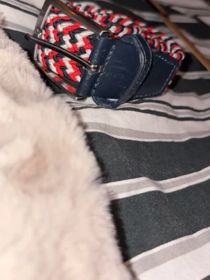 Randigt flätat bälte med blå detaljer - Snyggt flätat bälte med randigt mönster i rött, vitt och blått. Bältet har mörkblå läderdetaljer vid spännet och änden, samt ett klassiskt metallspänne. Perfekt för att ge outfiten en färgklick och funkar till många olika stilar. Bältet har några få skråmor men inget man märker.