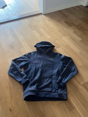 Mörkblå hoodie från Lyle & Scott - Snygg mörkblå hoodie från Lyle & Scott med klassisk gul logga på bröstet. Hoodien har huva, känguruficka och ribbade muddar vid ärmslut och nederkant. Perfekt för en chill och stilren look.