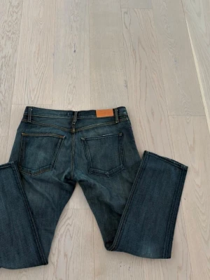 Mörkblå raka jeans från Acne - Säljer ett par mörkblå jeans från Acne med raka ben och klassisk femficksmodell. Jeansen har kontrastsömmar och en läderpatch bak i midjan. Materialet är slitstark denim i bomull, perfekt för en avslappnad och stilren look.