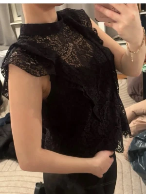 Svart spetsblus med volangdetaljer - Säljer en svart blus i spets med volangdetaljer och hög hals. Blusen är ärmlös och har ett transparent mönster upptill som ger en elegant look. Perfekt att styla med både jeans och kjol för en trendig outfit.