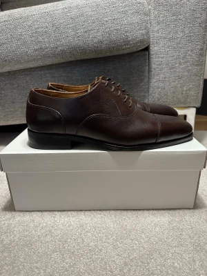 Mörkbruna Oxfordskor från These Glory Days - Nypris: 1599 kr. Säljer ett par OANVÄNDA mörkbruna läderskor från These Glory Days, handgjorda i Portugal! Skorna har klassisk snörning, låg klack och elegant rundad tå. Perfekta för dig som gillar tidlös och clean stil. OBS: Storlek 44, Små i storlek. 