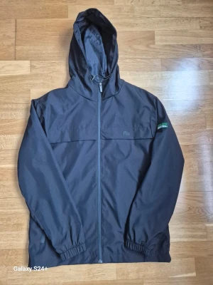 Svart vindjacka från Lacoste - Stilren svart vindjacka från Lacoste med huva och dragkedja framtill. Jackan har elastiska muddar vid ärmsluten och en liten Lacoste-logga på bröstet. Tillverkad i lätt och tåligt material. 