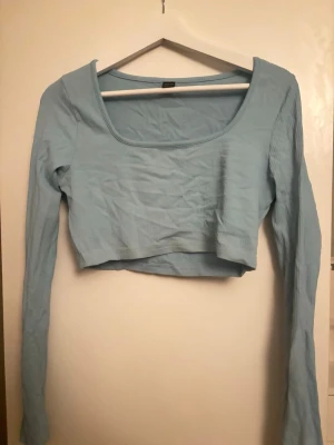 Ljusblå croppad långärmad topp - Trendig ljusblå croppad topp med lång ärm och bred, rundad urringning. Tillverkad i ett ribbat material som ger en snygg struktur. Perfekt att matcha med höga jeans eller kjol för en cool look.