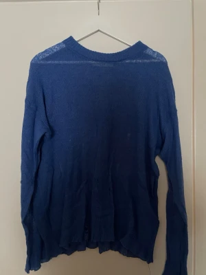 Blå tunn stickad tröja - En blå, tunnstickad tröja med lång ärm och avslappnad passform. Tröjan har en enkel rund halsringning och är tillverkad i ett lätt material som ger en luftig känsla. Perfekt för lager på lager och en chill look. Designen är med små hål här och var.