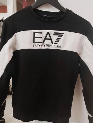Svartvit sweatshirt från Emporio Armani - Säljer en svartvit sweatshirt från Emporio Armani EA7 med stor logga på bröstet. Tröjan har en bred vit panel över bröstet och armarna, rund halsringning och är tillverkad i mjukt material. Perfekt för dig som gillar streetwear och stilrena detaljer.