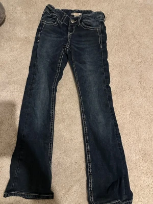 Mörkblå bootcut jeans med brodyr - Säljer ett par mörkblå bootcut jeans med kontrastsömmar och snygg brodyr på bakfickorna. Jeansen har låg midja och klassisk femficksdesign. Perfekta för dig som gillar detaljer och en avslappnad stil.