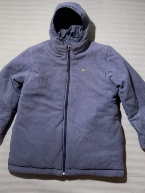 Ljuslila fleecejacka från Nike - Mysig ljuslila fleecejacka från Nike med huva och hel dragkedja framtill. Liten gul Nike-logga på bröstet och diskret broderad text på ryggen. Jackan har lång ärm och är tillverkad i mjuk fleece som håller dig varm under kyliga dagar.                             (NJ0004)