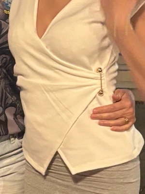 Vit omlottopp med gulddetaljer - Snygg vit omlottopp med v-ringning och dekorativa guldfärgade knappar i sidan. Toppen har en figurnära passform och är tillverkad i ett mjukt material som känns bekvämt mot huden. Perfekt för dig som vill ha en stilren och trendig look.