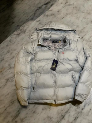 Ljusgrå dunjacka från Polo Ralph Lauren - Snygg ljusgrå dunjacka från Polo Ralph Lauren med klassisk puffer-design och röd logga på bröstet. Jackan har dragkedja framtill, två sidofickor med dragkedja och hög krage. Perfekt för kalla vinterdagar och riktigt stilren look. ⚠️ Är stor i storlek ⚠️ 