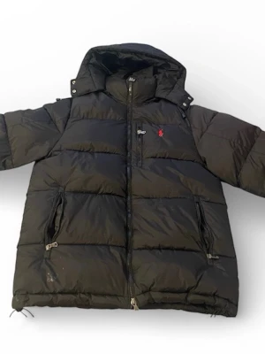 Svart pufferjacka från Polo Ralph Lauren - Säljer en svart pufferjacka från Polo Ralph Lauren med huva och dragkedja framtill. Jackan har två stora fickor med dragkedja och den klassiska röda Polo-loggan på bröstet. Perfekt för kalla vinterdagar och riktigt skön att bära.