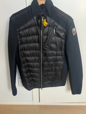 Svart quiltad pufferjacka Parajumpers - ÄKTA HELT NY PARAJUMPER CARDIGAN Svart pufferjacka från Parajumpers med quiltad framsida och rygg, samt släta ärmar. Jackan har dragkedja framtill, bröstficka med dragkedja och en gul detalj vid halsen. Märkespatch på ena ärmen och hög krage för extra stilpoäng.