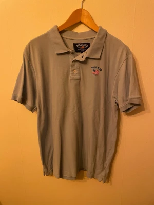 Ljusgrå piké från Polo Club - Snygg ljusgrå pikétröja från Polo Club med klassisk krage och två knappar framtill. Diskret broderad logga på bröstet med amerikansk flagga. Perfekt till jeans eller shorts för en clean och enkel stil.