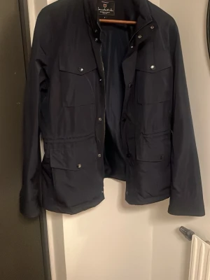 Bondelid Gerald Fieldjacket  - Stilren mörkblå jacka från Bondelid med fyra stora fickor framtill och hög krage. Jackan har dragkedja och tryckknappar samt en rak passform. Perfekt för dig som gillar en enkel men funktionell look.