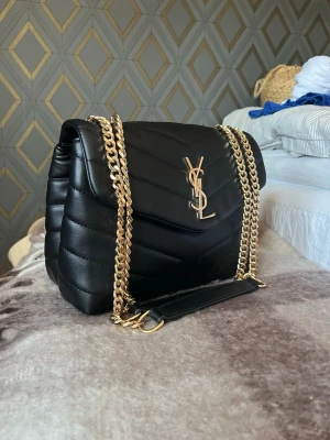 Svart YSL handväska med guldkedja - Snygg svart handväska från Saint Laurent i quiltat läder med ikonisk YSL-logga i guld framtill. Väskan har en tjock guldfärgad kedjerem och lockstängning. Perfekt storlek för det viktigaste och ger en lyxig touch till din outfit. (Ej äkta)