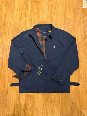  Polo Ralph Lauren Jacka navy blue, size L - Stilren marinblå jacka från Polo Ralph Lauren med klassisk krage och dragkedja. Insidan har ett rutigt foder i rött, grönt och gult. Jackan har justerbara band i midjan och knappar vid ärmsluten. Perfekt för dig som gillar tidlös och snygg design.