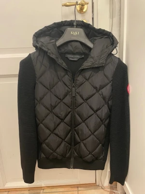 Svart cardigan från Canada Goose - Svart pufferjacka från Canada Goose med quiltat mönster på framsidan och huva. Ärmar och sidor är ribbstickade i tyg, medan framsidan och huvan är i glansigt material. Dragkedja framtill och röd logga på ärmen ger en clean och modern look. Storlek s men sitter som xs. Nästan oanvänd och har inga synliga defekter.