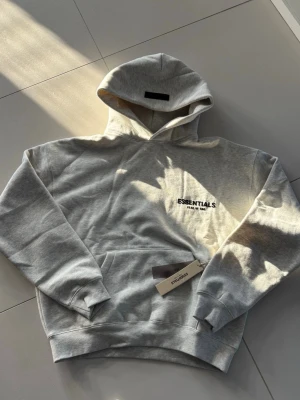 Grå Essentials Fear of God hoodie - Snygg grå hoodie från Essentials Fear of God med svart logga på bröstet. Klassisk känguruficka framtill och stor huva. Tillverkad i mjukt bomullsmaterial som känns riktigt skönt mot huden. Perfekt för dig som gillar streetwear och enkel stil.