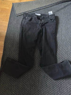 Svarta jeans från Mood Denim - Säljer ett par svarta jeans från Mood Denim i storlek 30/30. Jeansen har klassisk femficksmodell, normal midja och raka ben. Materialet är mjukt denimtyg som ger en avslappnad look. Perfekta för dig som gillar stilrena och enkla jeans.
