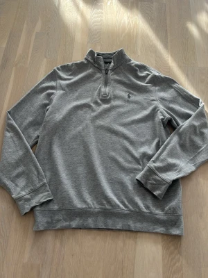 Grå halvzip tröja från Polo Ralph Lauren - Snygg grå långärmad tröja från Polo Ralph Lauren med halv dragkedja och liten broderad logga på bröstet. Perfekt för en avslappnad men stilren look.