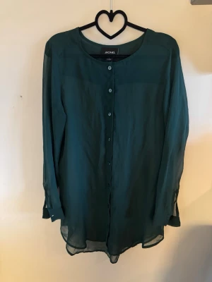 Mörkgrön transparent blus från MONKI - Stilren mörkgrön blus från MONKI i transparent, lätt material. Blusen har långa ärmar, rund halsringning och knäppning framtill med små knappar. Perfekt för lager på lager och en chill men ändå snygg look. 100% polyester.