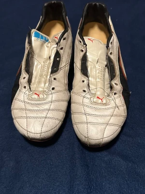 Puma Spirit vita fotbollsskor - Säljer ett par klassiska Puma Spirit fotbollsskor i vitt med svarta och röda detaljer. Skorna har en låg profil, sydda mönster på ovandelen och Puma-logga på tungan och sidan. Materialet är syntetläder och designen är sportig och retro. Snören medföljer 