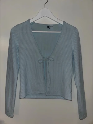 Ljusblå ribbad topp med knyt - Snygg ljusblå ribbad topp med v-ringning och knyt framtill. Toppen har långa ärmar och croppad passform, perfekt att styla med höga jeans eller kjol. Enkel men trendig design som passar till många olika looks. Har ett hål men går lätt att sy ihop.