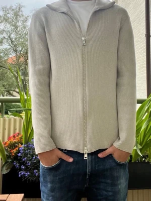 Grå ziptröja från Ljung By Marcus Larsson - Säljer en stilren beige ziptröja från Ljung. Tröjan är ribbstickad med hög krage och dragkedja framtill. Perfekt för lager på lager och enkel att matcha med olika outfits. Materialet känns mjukt och bekvämt mot huden.