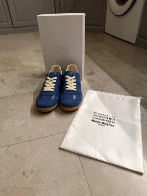 Blå Maison Margiela sneakers - Snygga blå sneakers från Maison Margiela med vita skosnören och beige detaljer. Skorna har en klassisk låg siluett och en gummisula som ger en retrovibe. Tillverkade i mocka och läder för en exklusiv känsla. Perfekta för dig som vill sticka ut med stil.