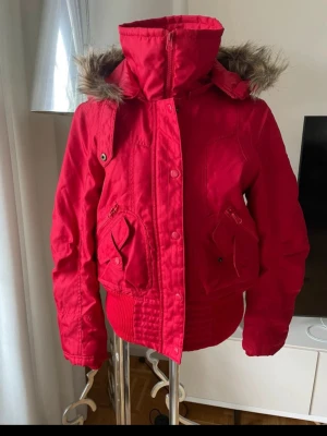Röd pufferjacka med fuskpäls - Snygg röd pufferjacka med stor huva och fuskpälsdetalj. Jackan har hög krage, två stora fickor framtill med dragkedja och knappar samt ribbade muddar i midja och ärmslut. Perfekt för kalla dagar och ger en cool streetwear-look.