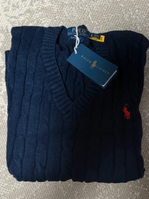 Mörkblå kabelstickad tröja Polo Ralph Lauren - Kabelstickad mörkblå tröja från Polo Ralph Lauren med V-ringning och klassisk röd logga broderad på bröstet. Tröjan har ribbade muddar och är tillverkad i mjukt material som passar perfekt till lager-på-lager-stil.