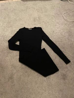 Svart långärmad klänning - En enkel och stilren svart klänning med lång ärm och rund halsringning. Klänningen är figurnära och har en kort längd, perfekt för dig som gillar minimalistisk stil. Materialet känns mjukt och stretchigt, vilket gör den bekväm att bära. Klänningen är ribbad och är från New Yorker.