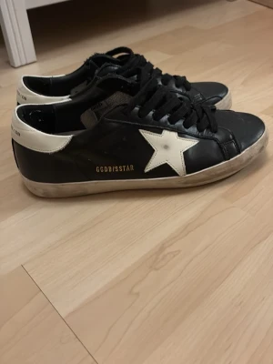 Svarta Golden goose sneakers med vit stjärna - Snygga svarta sneakers från Golden Goose med vit stjärndetalj på sidan och vit häl. Skorna har svarta skosnören och en slitstark sula. Klassisk låg modell i läder som ger en cool och avslappnad vibe. Perfekta för dig som gillar streetstyle och unika detaljer. Storlek 44. Skoldåd ingår
