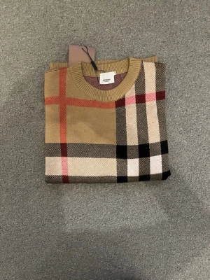Beige rutig tröja från Burberry - Snygg beige tröja från Burberry med klassiskt rutigt mönster i svart, vitt och rött. Tröjan har rund halsringning, ribbade muddar och är långärmad. Perfekt för dig som gillar stilrena och tidlösa plagg.