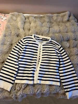 Randig kofta med knappar - Snygg kofta med marinblå och vita horisontella ränder. Rund halsringning och knäppning framtill med en guldig knapp . Mjuk och tunn stickning, perfekt för lager på lager. Klassisk design som passar till det mesta! 