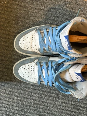 Nike Air Jordan 1 hyper royal  High blå/vit - Snygga Nike Air Jordan 1 Hyper Royal High sneakers i vitt och ljusblått läder med grå swoosh och blå skosnören. Klassisk high-top siluett med perforerad tåbox och ikonisk Air Jordan-logga på sidan. Perfekta för dig som gillar streetwear och retrostil.kommer även Med låda från restock och extra skosnören