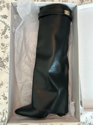 Svarta höga boots med spetsig tå - Säljer ett par svarta höga stövlar i slätt skinnimitation med spetsig tå och bred skaft. Stövlarna har en dekorativ rem med guldfärgat spänne upptill och en diskret kilklack. Perfekta för en edgy och trendig look. !!Använt dem en gång!!