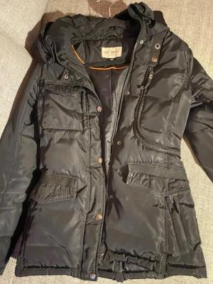 Svart parkasjacka från Roco Baroco - Svart parkasjacka med huva från Roca Baroco. Jackan har flera praktiska fickor med lock och dragkedja, knappar och dragkedja framtill samt justerbar midja. Tillverkad i ett slitstarkt, vadderat material som håller värmen under kalla dagar.