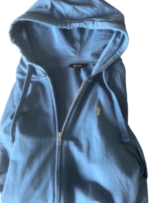 Blå zip hoodie från Next - Blå hoodie med dragkedja från Next. Har huva med dragsko, fickor framtill och ett litet gult broderat motiv på bröstet. Tillverkad i mjukt bomullsmaterial som är skönt att ha på sig. Perfekt för en avslappnad och chill stil.