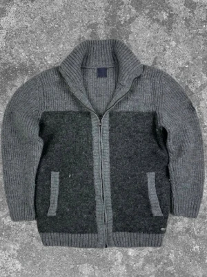 Mörkgrå stickad kofta med dragkedja - Mysig mörkgrå stickad kofta med hög krage och dragkedja framtill. Koftan har två fickor och ribbstickade detaljer vid ärmslut, nederkant och krage. Perfekt för lager på lager och en avslappnad stil.